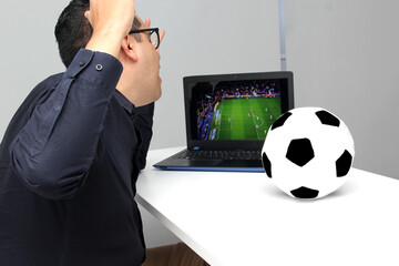 Latino adult man watches a World Cup soccer game on his laptop in his office while working next to a soccer ball during work hours in the morning
