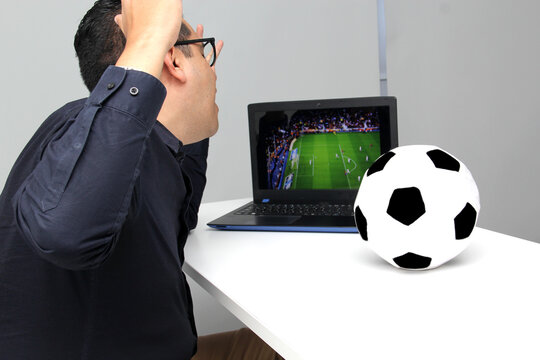 Latino adult man watches a World Cup soccer game on his laptop in his office while working next to a soccer ball during work hours in the morning