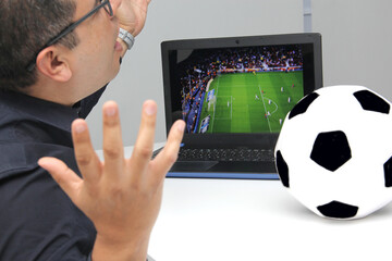 Latino adult man watches a World Cup soccer game on his laptop in his office while working next to a soccer ball during work hours in the morning 