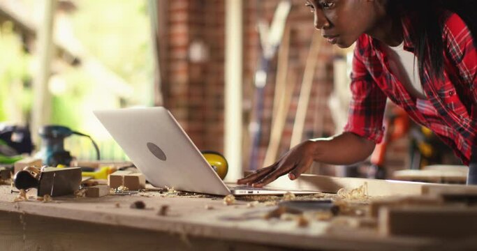 African American female carpenter working with computer laptop in workshop . craftsman wearing safety glasses checking order of clients or learning online