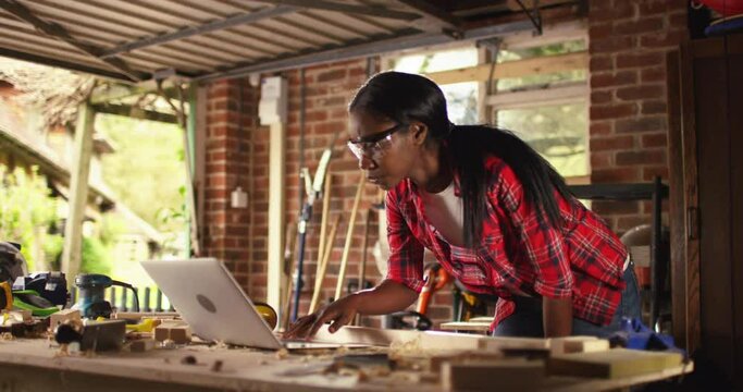 African American female carpenter working with computer laptop in workshop . craftsman wearing safety glasses checking order of clients or learning online