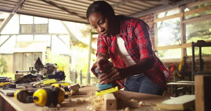 African American female carpenter wearing protective mask sanding a wooden plank with electric sander. carpentry, craftsmanship and handwork concept