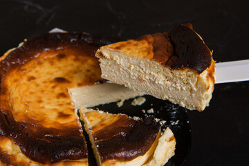 A piece of Basque cheesecake is raised on a knife above a dessert cut into pieces, selective focus