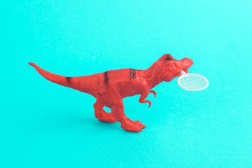 Toy red dinosaur tyrannosaurus rex with tennis racquet on turquoise background. Minimalism creative...
