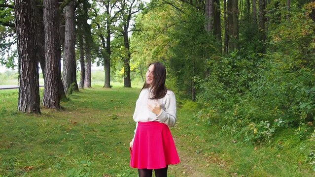 Young girl puts on red hat on her head and corrects long dark hair. Beautiful woman in gray sweater and red skirt posing in the forest on a sunny autumn day. Walking and relaxing in the woods.