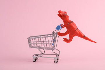Toy red dinosaur tyrannosaurus rex with supermarket trolley on pink background. Minimalism creative...