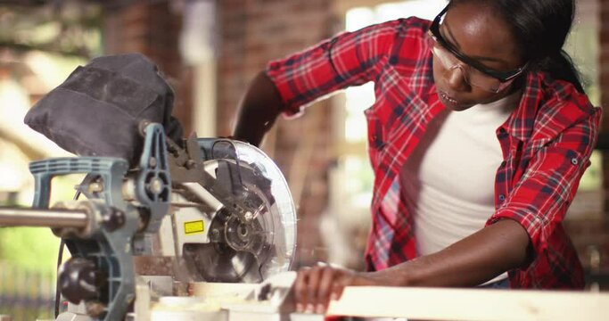 African American female carpenter cutting wooden plank with a circular electric saw in carpentry workshop