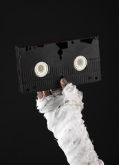 Mummy's hand wrapped in bandage holds retro vhs video cassette isolated on black background....