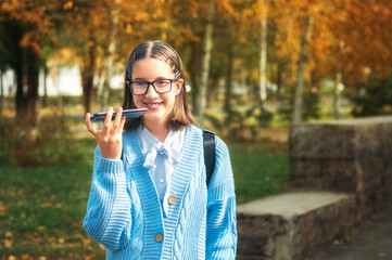 A young schoolgirl with a backpack is walking down the street on a sunny autumn day and talking on the phone.