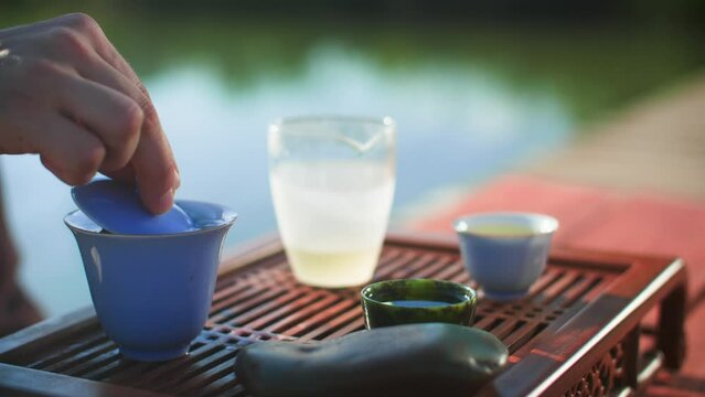 Closeup of man checking tea leaves in gaiwan container at chaban wooden table. Male is making Oolong green tea for Chinese tea ceremony on pier amidst lake. He is enjoying refreshment during summer.