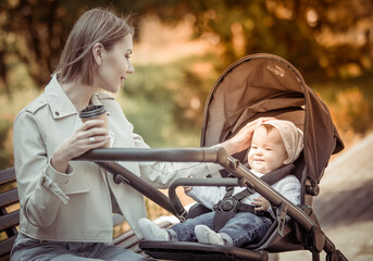 Beautiful Caucasian mother sits on a bench with baby in stroller outdoor