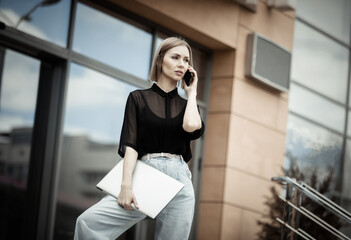 Fototapeta premium Modern business lady talking on the phone and holding laptop on the stairs outdoors