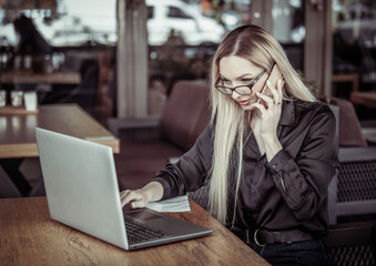 Attractive blonde business woman talking on phone and using laptop while sitting at table in outdoor cafe