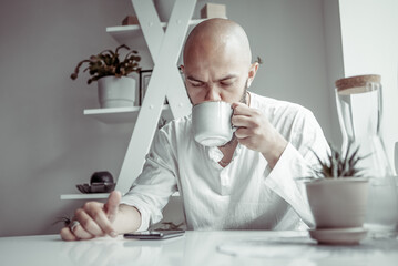 Handsome bald bearded man drinking coffee and using smartphone while sitting at the table in modern...