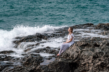 A woman in a storm sits on a stone in the sea. Dressed in a white long dress, waves crash against the rocks and white spray rises.