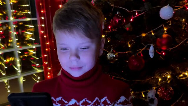 A cute smiling boy looks at the phone in the evning in front of the Christmas tree. leisure, technology and holidays concept