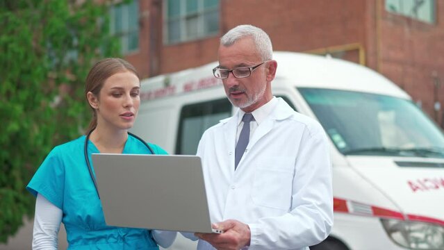 Close Up Of Caucasian Young Professional Woman Nurse Looking At Laptop And Consulting With Experienced Man Doctor Standing Outdoor Near Ambulance. Hospital Workers In Uniform Discussing Treatment