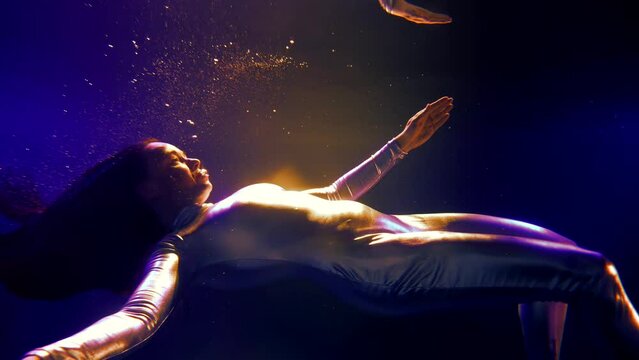 a woman emerges from under the water she is in a silver jumpsuit like a mermaid swimming in neon lights
