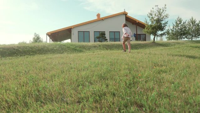 Caucasian cute ittle boy playing with toy plane running outdoor near home. Positive small child plays in green field near camp house on sunny day and smiling. Cheerful kid with toy. Nature, childhood