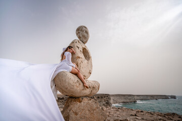 A woman sits on a stone sculpture made of large stones. She is dressed in a white long dress,...
