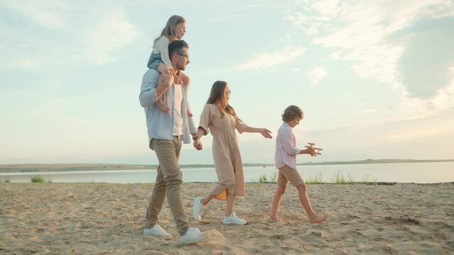 Caucasian family with little kids walking near water in nature. Happy parents with children walk on a beach near beautiful lake. Recreation. Family rest. Boy playing with toy plane. Happiness concept