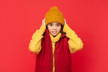 kid in knitted winter hat and down vest feel cold on red background, fashion