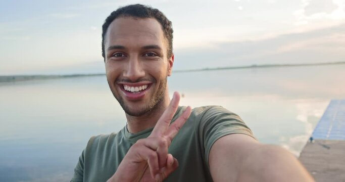 Close up of joyful young man athlete in good mood standing on lake posing to smartphone camera and smiling. POV of African American happy male taking selfie photos on cellphone outdoors