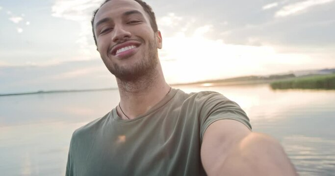 POV of joyful young man athlete in good mood standing on lake posing to smartphone camera smiling and showing V sign on sunset recording video. African American happy guy taking selfie photo on gadget