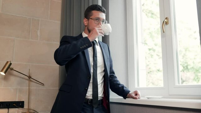 Portrait of Caucasian young businessman in suit standing indoors near window holding cup of coffee in hands. Business concept. Handsome man drinking coffee in room looking in window.