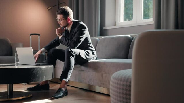 Caucasian handsome adult businessman sitting on sofa in hotel lobby typing on laptop working online. Business trip concept. Stylish elegant male entrepreneur traveling for work. Technology device