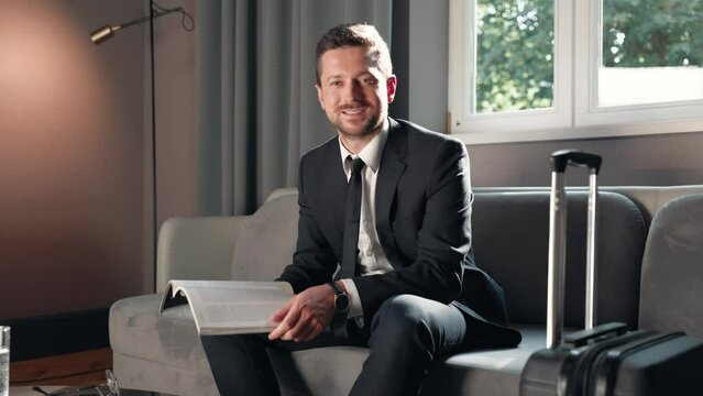 Handsome cheerful Caucasian businessman eading journal while waiting for business meeting sitting on sofa in hotel lobby smiling to camera. Business people concept. Guy in suit reading magazine