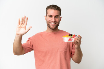 Young caucasian man holding a bowl of fruit isolated on white background saluting with hand with happy expression