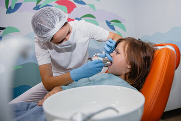 A woman dentist in a white coat and a medical cap treats the teeth of European appearance girl in a dental office