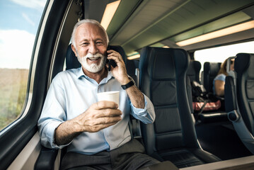 Smiling businessman talking on smart phone while commuting and having coffee on a railroad train.