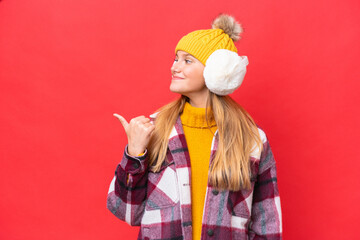 Young beautiful woman wearing winter muffs isolated on red background pointing to the side to present a product