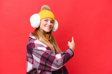 Young beautiful woman wearing winter muffs isolated on red background pointing back