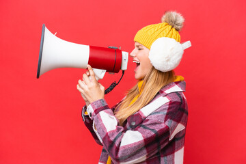 Young beautiful woman wearing winter muffs isolated on red background shouting through a megaphone