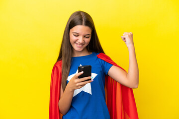 Little caucasian superhero girl isolated on yellow background with phone in victory position © luismolinero
