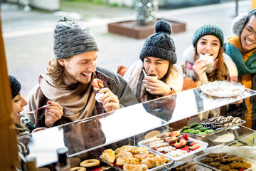 Happy friends having fun eating at Christmas market on winter holidays - Urban vacation concept with young people hanging out together by candy shop