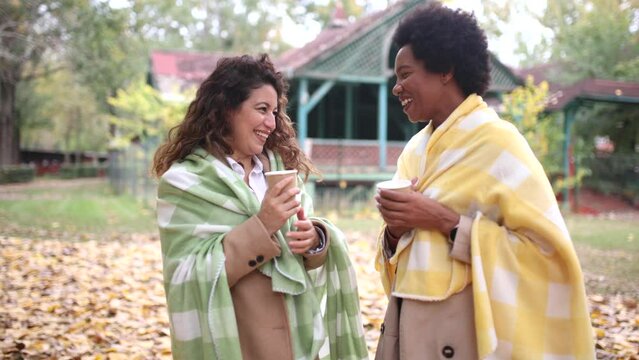 Two smiling young women friends chatting outdoors and drink coffee while enjoying walk in park together