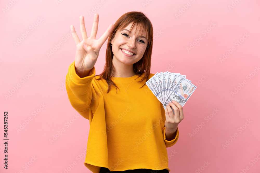 Wall mural redhead girl taking a lot of money isolated on pink background happy and counting four with fingers