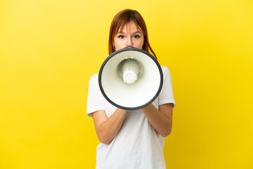Redhead girl isolated on yellow background shouting through a megaphone to announce something