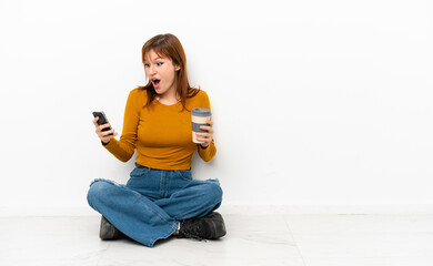 Redhead girl sitting on the floor isolated on white background holding coffee to take away and a mobile