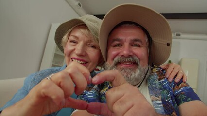 Portrait of carefree smiling retired senior traveler couple in sunhats posing for selfie shot on smartphone, showing heart shape with hands, expressing excitement and happiness indoors. - Powered by Adobe
