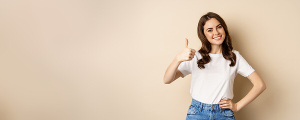 Portrait of modern young woman showing thumbs up, like and approve, smiling pleased, recommending company or website, standing over beige background