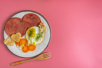 Breakfast plate with fresh carrots, miniature pancakes in the form of heart, sunny side up egg, roasted Bologna sausage over pink background. Free space for text.