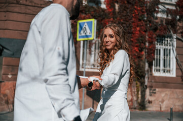 Fototapeta premium Having fun and holding each other by hands. Beautiful young couple is together on the street against old building