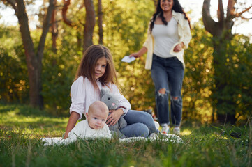 Fototapeta premium Kid is sitting with her little brother on the ground with mother behind. In the forest. Having weekend