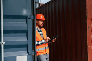 Male worker is on the location with containers. Using digital tablet