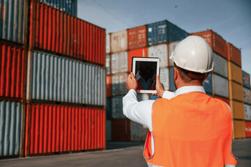 Holding tablet and making photo of the objects. Male worker is on the location with containers